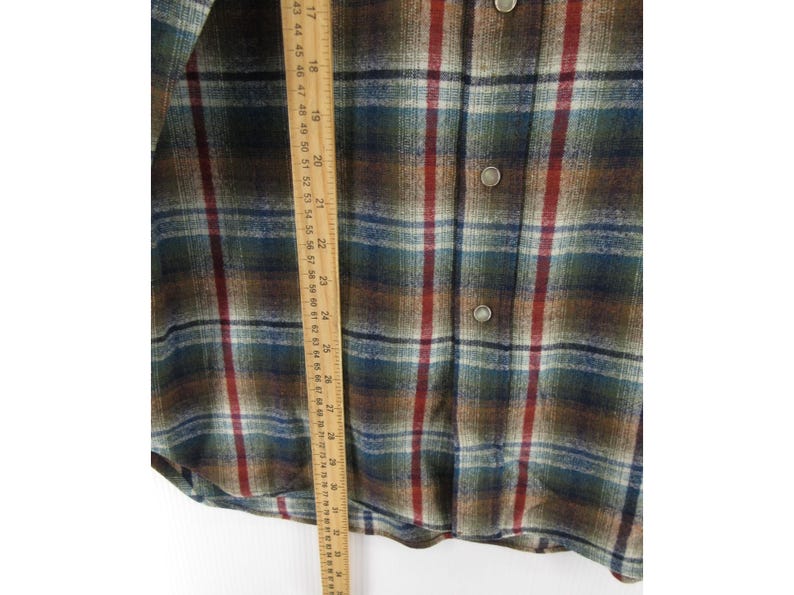 May include: A long-sleeve button-down shirt featuring a plaid pattern in shades of brown, dark green, blue, and red. A wooden ruler is placed vertically over the fabric, showing measurements in inches and centimetres for scale and reference.