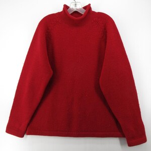 Könnte beinhalten: Ein roter Rollkragenpullover mit langen Ärmeln. Der Pullover besteht aus einem strukturierten Strickstoff und hängt an einem weißen Kleiderbügel. Ein klassischer Stil, geeignet für kühleres Wetter.