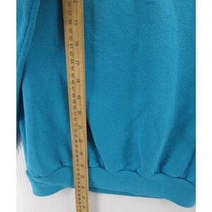 May include: A solid teal sweatshirt featuring a ribbed hem and cuffs, positioned next to a wooden ruler that shows a length measurement extending from approximately 14 inches to 26 inches along the side of the garment fabric.