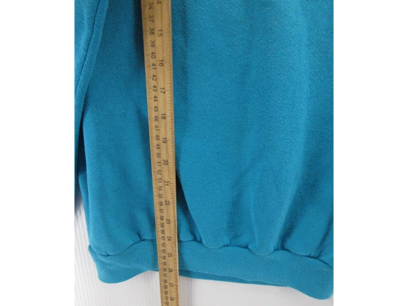 May include: A solid teal sweatshirt featuring a ribbed hem and cuffs, positioned next to a wooden ruler that shows a length measurement extending from approximately 14 inches to 26 inches along the side of the garment fabric.