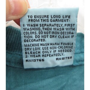 May include: A light blue garment tag with black text. The tag provides washing instructions: wash separately, wash with colors, do not iron, do not dry clean if decorated. Machine wash warm, tumble dry low, use non-chlorine bleach. Wear repeatedly.