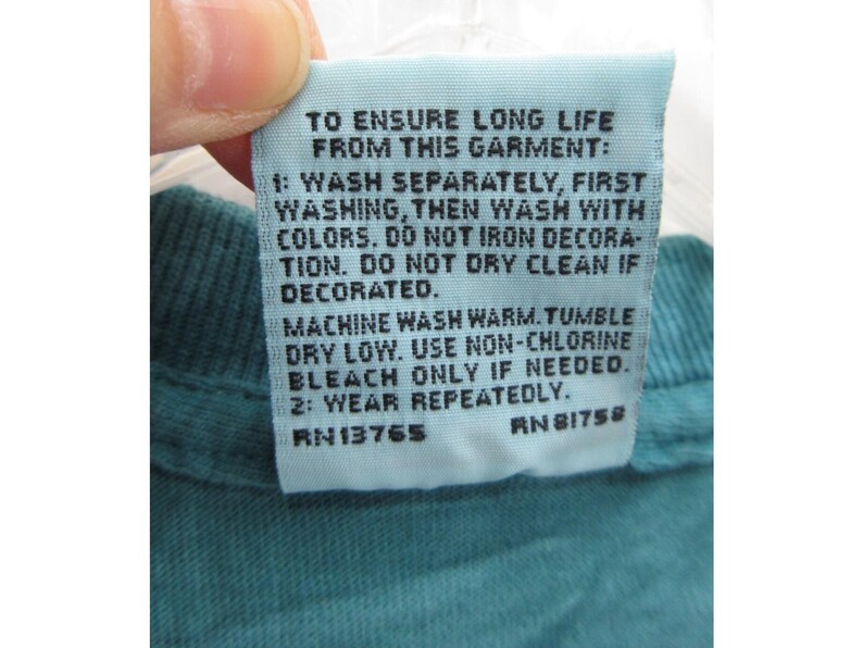 May include: A light blue garment tag with black text. The tag provides washing instructions: wash separately, wash with colors, do not iron, do not dry clean if decorated. Machine wash warm, tumble dry low, use non-chlorine bleach. Wear repeatedly.