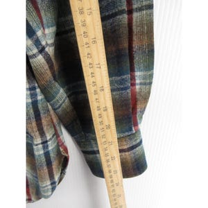 May include: A long-sleeved plaid shirt featuring a pattern of green, blue, brown and red lines. A wooden ruler is placed vertically against the sleeve, showing a measurement length extending from approximately 38 cm to 64 cm in total.