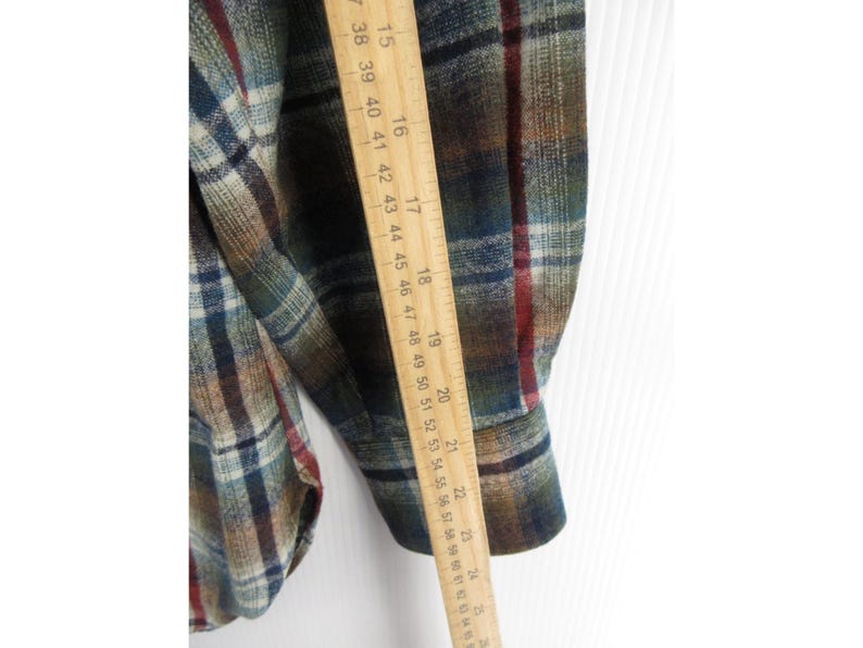 May include: A long-sleeved plaid shirt featuring a pattern of green, blue, brown and red lines. A wooden ruler is placed vertically against the sleeve, showing a measurement length extending from approximately 38 cm to 64 cm in total.