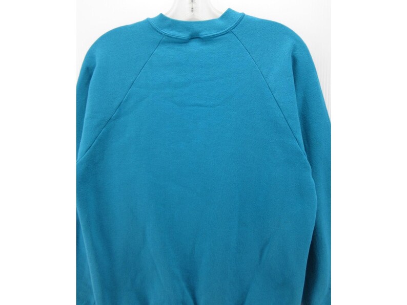 May include: A solid teal sweatshirt featuring a crew neckline and long raglan sleeves. The garment is constructed with a ribbed collar and cuffs, showcasing a plain, unpatterned design throughout the front and back of the fabric.