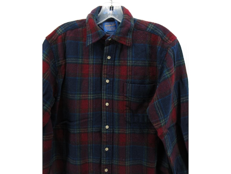 May include: A long-sleeve button-down shirt featuring a plaid pattern in shades of dark red, navy blue, and thin green lines. The shirt includes a standard collar, a single chest pocket on the left side, and light-colored buttons down the front placket.