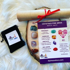 May include: A set of crystals for the zodiac sign Aries, with a card listing the crystals and their properties. The crystals are Amazonite, Carnelian, Amethyst, and Red Jasper. The card also includes the astrological symbol for Aries and a description of the Aries personality. The card is on a wooden base with a brown paper scroll and a black velvet bag.