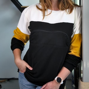 K&ouml;nnte beinhalten: Ein lang&auml;rmliges Shirt in Schwarz, Wei&szlig; und Gelb mit einem Mesh-Panel auf der Vorderseite. Das Shirt hat eine Rei&szlig;verschlusstasche auf der Brust. Die Person tr&auml;gt eine wei&szlig;e Uhr mit schwarzem Zifferblatt.