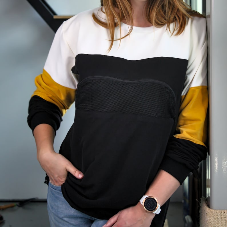 K&ouml;nnte beinhalten: Ein lang&auml;rmliges Shirt in Schwarz, Wei&szlig; und Gelb mit einem Mesh-Panel auf der Vorderseite. Das Shirt hat eine Rei&szlig;verschlusstasche auf der Brust. Die Person tr&auml;gt eine wei&szlig;e Uhr mit schwarzem Zifferblatt.