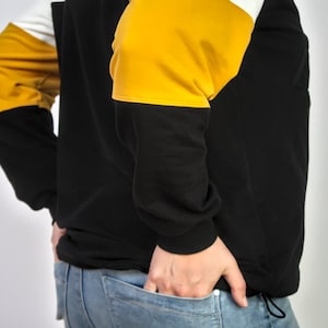 K&ouml;nnte beinhalten: Ein schwarzer Pullover mit gelben und wei&szlig;en Farbfeldern. Der Pullover wird von einer Person getragen, die ihre H&auml;nde in den Taschen hat. Die Person tr&auml;gt eine blaue Jeans.