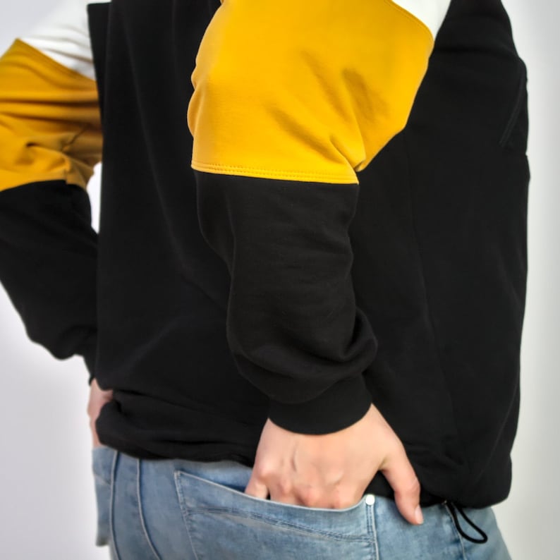 K&ouml;nnte beinhalten: Ein schwarzer Pullover mit gelben und wei&szlig;en Farbfeldern. Der Pullover wird von einer Person getragen, die ihre H&auml;nde in den Taschen hat. Die Person tr&auml;gt eine blaue Jeans.