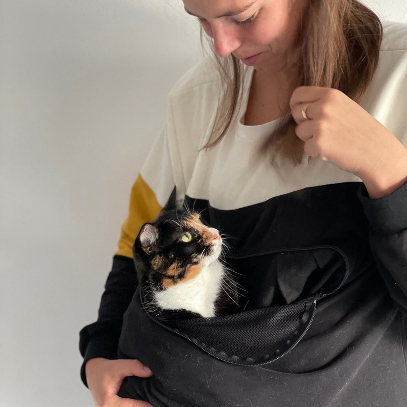 K&ouml;nnte beinhalten: Eine Frau tr&auml;gt ein schwarz-wei&szlig; gestreiftes Shirt mit einem gelben Streifen am &Auml;rmel und h&auml;lt eine dreifarbige Katze in einer schwarzen Tasche mit einem Netzfenster. Die Katze schaut die Frau an.