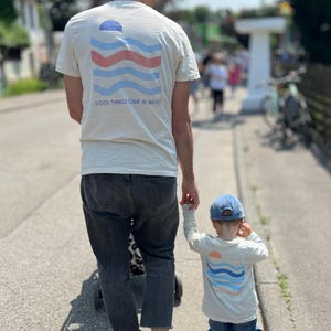 Könnte beinhalten: Ein Erwachsener und ein Kind gehen einen Weg entlang, beide tragen cremefarbene T-Shirts mit einem Wellenmuster und dem Text "GOOD THINGS COME IN WAVES". Das Kind trägt eine blaue Kappe und Jeans.