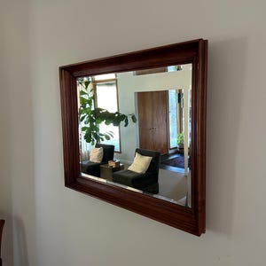 May include: A rectangular wall mirror featuring a polished, dark brown wooden frame with tiered, beveled edges. The glass surface is clear and reflects a room interior containing two dark chairs with white cushions and a green plant.