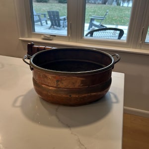 May include: A copper-colored metal pot with two small handles. The pot has a rounded shape with a slightly flared rim and a dark interior. The pot is sitting on a white countertop.