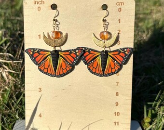 Könnte beinhalten: Ein Paar goldene Halbmond-Ohrringe mit orangefarbenen Glasperlen und einem Monarchfalter-Design. Die Ohrringe werden auf einem Holzlineal mit Zoll- und Zentimetermarkierungen gezeigt.