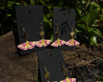 Könnte beinhalten: Ohrringe mit einem Motten-Design, mit rosa und gelben Flügeln, goldfarbenen Sonnenstrahl-Akzenten und goldfarbenen Haken. Die Ohrringe werden auf schwarzen Ohrringkarten präsentiert. Der Hintergrund umfasst Grün und ein Stück Holz.