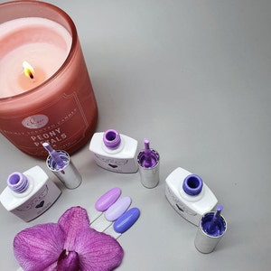 May include: Four bottles of nail polish in shades of purple and lavender. The bottles are labeled "Angel" and have a heart design. The bottles are arranged on a gray surface with a pink orchid and a candle.