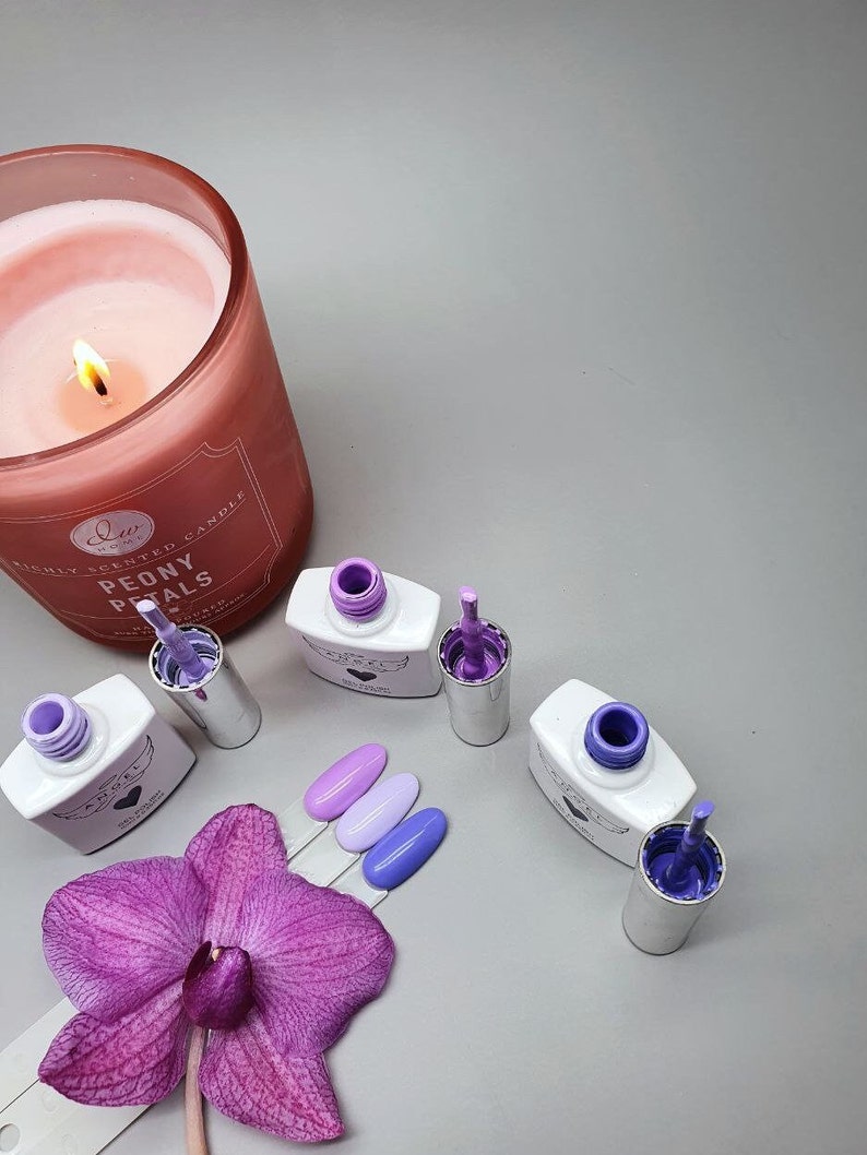 May include: Four bottles of nail polish in shades of purple and lavender. The bottles are labeled "Angel" and have a heart design. The bottles are arranged on a gray surface with a pink orchid and a candle.