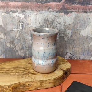 Könnte beinhalten: Eine kleine, hellrosa Keramikvase mit einem gesprenkelten Überzug und blauen Akzenten. Die Vase steht auf einer Holzoberfläche.