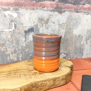 May include: A small, orange and grey ceramic cup with a textured finish. The cup is sitting on a wooden chopping board.