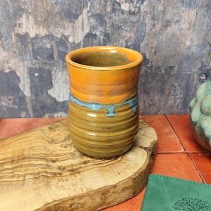 Op de afbeelding: Een bruine keramische mok met een lichtblauwe glazuur en een gele glazuur aan de bovenkant. De mok staat op een houten snijplank.