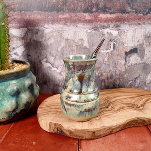 May include: A ceramic mate gourd with a glossy, mottled glaze in shades of teal, cream, and brown, featuring a flared rim and a narrow neck. A slender, metallic silver bombilla straw is inserted into the top of the vessel, which rests on a wooden board.