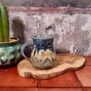 May include: A handcrafted ceramic mug with a blue and tan glaze rests on a wooden board. A green cactus in a turquoise pot is to the left. The background is a textured wall with red tile flooring.