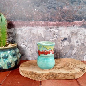 Puede incluir: Una pequeña taza de cerámica con acabado brillante que presenta una base turquesa, una banda central roja y un borde amarillo con un patrón de esmalte que gotea. La taza está colocada junto a un cactus verde en una maceta turquesa texturizada.
