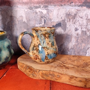 May include: A handcrafted ceramic mug with a distinctive glaze. The mug has a tan base with blue and brown splashes, and a curved handle. It rests on a wooden board, with a cactus in a blue pot to the left. The background is a textured wall.