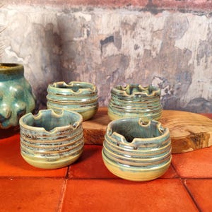 May include: Four small, handmade ceramic ashtrays with a ribbed design and a turquoise and brown glaze. The ashtrays are arranged on a wooden surface and a red-orange tile surface. A cactus in a turquoise pot is in the background.