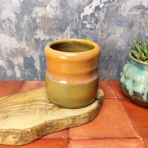 Puede incluir: Una taza pequeña de cerámica con un esmalte de dos tonos. La mitad superior de la taza es de color naranja claro, mientras que la mitad inferior es de color marrón oscuro. La taza está sentada sobre una superficie de madera.