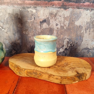 Può includere: Un bicchiere in ceramica fatto a mano con bordo azzurro e base gialla. La tazza è appoggiata su un tagliere ovale in legno. Lo sfondo presenta un muro strutturato con sfumature di marrone, rosso e grigio.