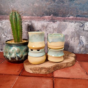 Puede incluir: Conjunto de macetas de cerámica hechas a mano en tonos azules y beige, apiladas sobre una tabla de madera. Un cactus en una maceta turquesa a la izquierda. Las macetas tienen una forma orgánica, ideales para suculentas. Altura aproximada: 10 cm.