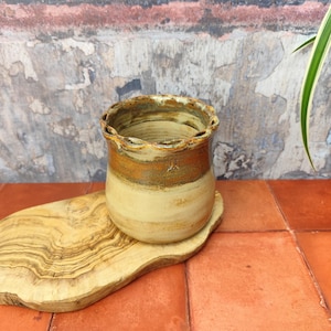 May include: A small, handmade ceramic planter with a scalloped rim. The planter is glazed in shades of brown, beige, and white. It is sitting on a wooden chopping board.