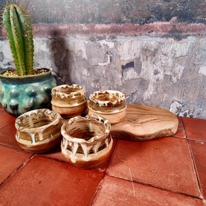 Puede incluir: Cuatro tazas de cerámica pequeñas y redondas con un esmalte bronceado y marrón que presenta patrones de goteo blanco en los bordes superiores. Las tazas están dispuestas sobre una tabla de madera junto a un cactus verde en una maceta azul verdosa.