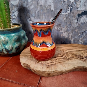 Può includere: Una zucca di yerba mate in ceramica fatta a mano con un design colorato. La zucca presenta una base rossa, che sfuma in arancione e giallo, con accenti blu sul bordo. Una cannuccia di metallo è all'interno. La zucca è appoggiata su un tagliere di legno.