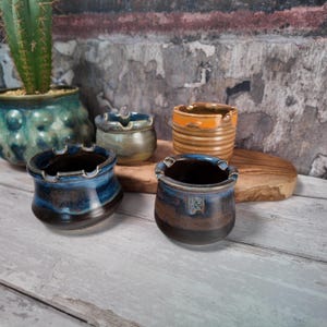 May include: Four small ceramic bowls with notched rims, featuring varied glazes in deep blue, brown, muted green, and ribbed orange. These vessels are arranged alongside a green cactus in a textured teal pot, all resting on a wooden board.
