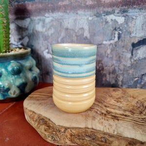 May include: A cylindrical ceramic tumbler featuring a two-tone glaze design. The upper portion is a soft teal blue with a smooth finish, while the lower section is a warm cream yellow with horizontal ribbed textures encircling the base.