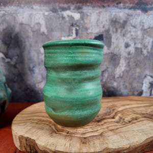 Può includere: Un bicchiere in ceramica verde fatto a mano con un design strutturato e scanalato. La tazza ha una base arrotondata e un bordo leggermente svasato. La glassa presenta variazioni di colore, creando un aspetto artigianale unico. Il bicchiere è esposto su una superficie di legno.