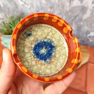 Könnte beinhalten: Ein kleiner, runder Aschenbecher aus Keramik mit einem blauen und weißen Mosaikdesign im Inneren und einem roten Rand mit orangefarbenen Punkten.