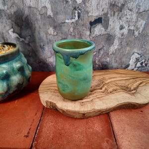 Könnte beinhalten: Ein handgefertigter Keramikbecher mit grüner und blauer Glasur. Der Becher hat eine zylindrische Form mit einem leicht ausgestellten Rand. Die Glasur hat einen Marmoreffekt mit Grüntönen und Blau. Der Becher steht auf einem Holzschneidebrett.