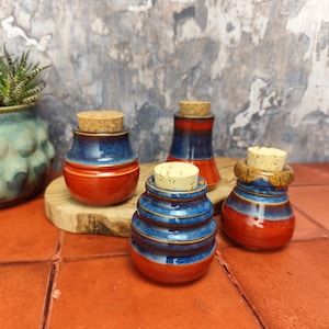 Op de afbeelding: Vier kleine keramische potten met kurken deksels. De potten zijn versierd met blauwe en oranje glazuur. De potten zijn gerangschikt op een houten oppervlak.