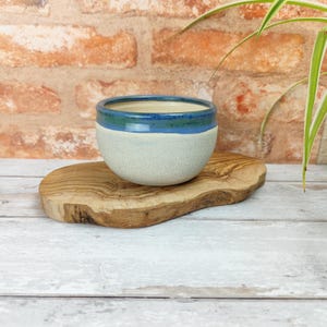 Op de afbeelding: Een kleine, witte keramische kom met een blauwe en groene rand. De kom staat op een houten snijplank.