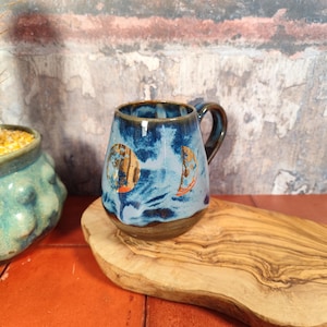 May include: A handcrafted ceramic mug, glazed in blue with gold moon phase patterns. It features a brown base and handle. The mug rests on a wooden chopping board. A unique piece of drinkware, perfect for hot beverages.