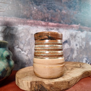 Puede incluir: Un jarrón de cerámica hecho a mano con forma cilíndrica. Presenta bandas horizontales de colores marrón, blanco y beige. El borde superior es de color marrón oscuro con detalles en negro. El jarrón está sobre una tabla de cortar de madera.