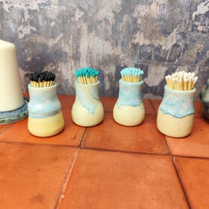 May include: Four ceramic matchstick holders with a light blue and yellow glaze. Each holder is filled with matches with different coloured tips: black, teal, teal, and white.