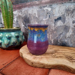 Op de afbeelding: Een handgemaakte keramische vaas met een levendig kleurenschema. De vaas heeft een dieppaarse basis, die overgaat in blauw en goud bij de rand. De vaas wordt gepresenteerd op een houten plank, met een cactus en een turquoise pot op de achtergrond.