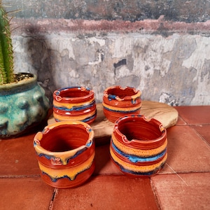 Puede incluir: Cuatro ceniceros cilíndricos pequeños de cerámica con acabado brillante. Cada pieza presenta una paleta de colores vibrantes en rojo intenso, naranja brillante y azul cobalto, con bandas horizontales y patrones de esmalte goteado.