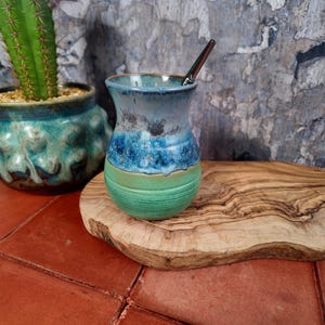Può includere: Una zucca di yerba mate in ceramica fatta a mano con una cannuccia in acciaio inossidabile. La zucca presenta una sfumatura di colori blu e verdi. Un cactus in un vaso turchese è sullo sfondo.